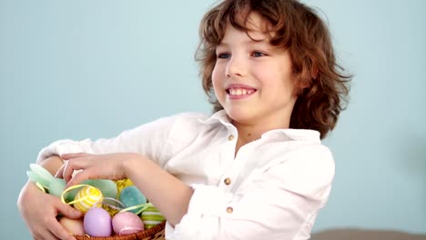 Niño-pelirrojo-rizado-en-una-camisa-blanca-con-una-figura-de-un-conejito-de-Pascua-y-un-conjunto-de-huevos-de-Pascua-en-una-cesta.-Un-niño-se-ríe-alegremente.-Cámara-lenta.