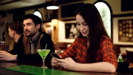 Junge-Schülerin-nutzt-Smartphone-sitzen-in-schicke-Bar-mit-Cocktails.-Sie-ist-Bildschirm-berühren-und-lächelnd.-Moderne-Wege-der-Kommunikationskonzept.