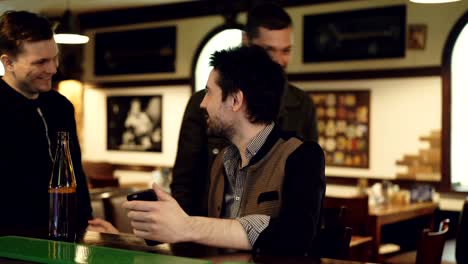 Young-handome-man-is-using-smartphone-at-bar-counter-when-his-male-friends-are-coming,-watching-screen-and-discussing-something.-Technology-communication-concept