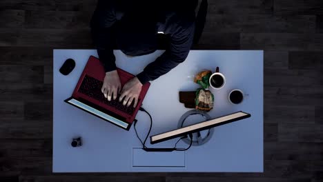 Young-man-in-black-hoodie-hacking-laptop-during-night,-sitting-behind-desk-with-laptop-and-monitor,-top-shot