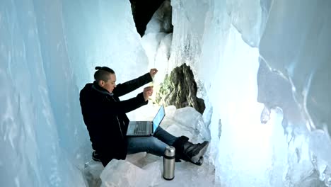 Man-is-sit-on-the-internet-in-laptop-in-an-ice-cave.-Around-the-mysterious-beautiful-ice-grotto.-User-communicates-in-social-networks.-He-crazy-strange-hipster-in-black-coat-and-fashionable-hairdo.