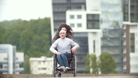 Feliz-discapacitado-en-silla-de-ruedas-con-mujer-joven-feliz-correr-en-la-calle-de-la-ciudad