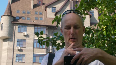 Bald-man-editing-video-on-smartphone-in-city-with-tree-and-building