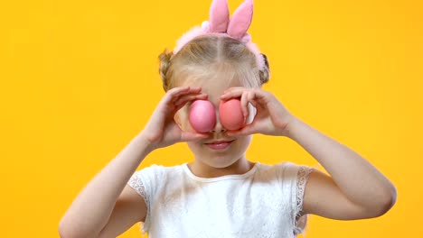 Cute-girl-having-fun-putting-colored-egg-to-eyes-on-yellow-background,-childhood