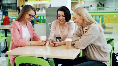 Happy-friends-good-looking-women-are-looking-at-smartphone-screen-and-talking-sitting-in-cafe-then-doing-high-five-and-laughing.-Modern-youth,-devices-and-friendship-concept.