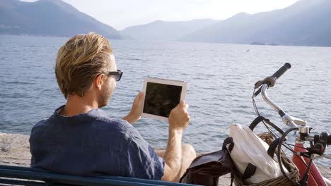 Young-man-using-digital-tablet-in-Swiss-village-near-lake-and-mountains--sitting-on-bench-with-bicycle-beside-him.-People-travel-technology-portable-devices-concept