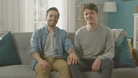 Pareja-Gay-masculino-atractivo-lindo-sentarse-juntos-en-un-sofá-en-casa.-Novio-pone-su-mano-en-pareja.-Ellos-son-feliz-y-sonriente.-Son-casualmente-vestida-y-su-habitación-tiene-un-Interior-moderno.