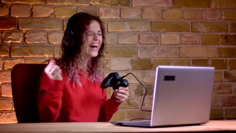 Portrait-of-young-female-blogger-in-red-hoodie-happily-playing-video-game-using-joystick-on-bricken-wall-background.