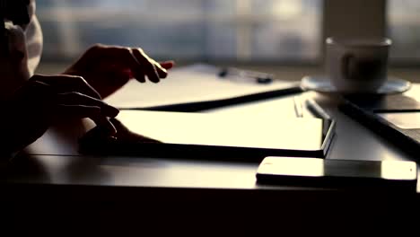 closeup,-dark-silhouette-of-female-hands.-she-is-typing-something-in-tablet,-next-is-a-cup,-mobile,-laptop-on-table.-Blurred-window-background-,-a-ray-of-light-is-reflected-in-the-tablet-screen