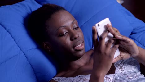 Smiling-black-american-woman-surfing-net-with-cellphone-at-home,lying-on-bed