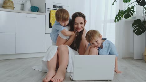 Moderne-Wohnung-liebende-Mutter-und-zwei-kleine-Söhne,-die-auf-dem-Boden-im-Wohnzimmer-sitzen,-schauen-auf-den-Laptop-Bildschirm.-Kinder-mit-Mama-spielen-auf-einem-Laptop