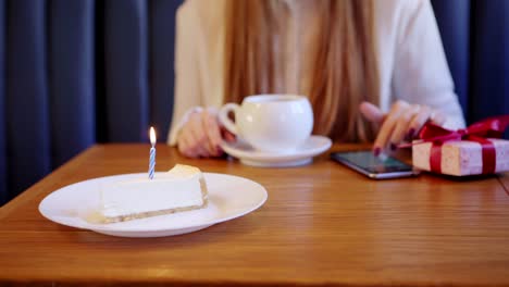 Close-up-de-niña-irreconocible-navegando-teléfono-inteligente-mientras-espera-a-su-amigo-en-la-mesa-en-la-cafetería-para-felicitar-en-el-cumpleaños.-Caja-de-regalo-y-tarta-de-queso-con-vela-ardiente-preparada-para-amigo.