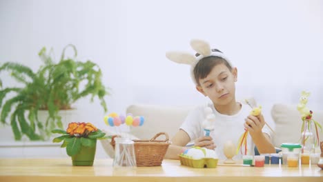 Young-adorable-boy-is-sitting-at-the-table-full-of-Easter-decorations-and-is-playing-with-Easter-bunnies-in-his-hands.-Bunnies'-discussion.-Who-would-paint-an-egg-Bunny-theatre.