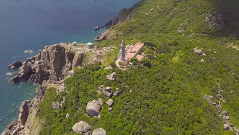 Luftblick-auf-den-Leuchtturm-des-Meeres-auf-grünem-Berg-und-felsigem-Ufer.-Schönes-Landschaftslichthaus-an-Felsen-und-Meeresufer-mit-blauer-Wasser-Drohne