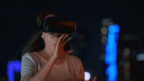 Junge-Frau-in-Virtual-Reality-Brille-in-der-Nachtstadt-bewegt-ihre-Hände