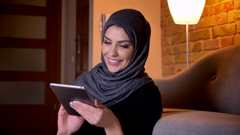 Nahauf-Porträt-von-erwachsenen-attraktiven-muslimischen-Frauen-im-Hijab-Surf-Web-auf-dem-Tablet-und-lächelnd,-während-auf-dem-Boden-in-der-Tür-in-einer-gemütlichen-Wohnung-sitzen