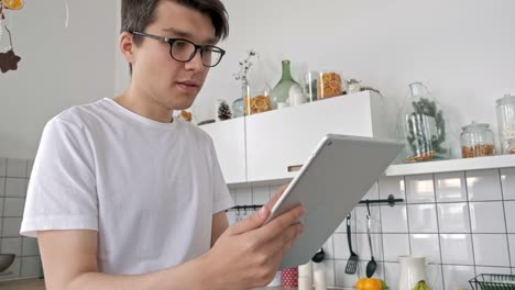 Attractive-man-at-home-using-tablet-in-kitchen-sending-message-on-social-media-smiling-enjoying-modern-lifestyle-wearing-white-shirt