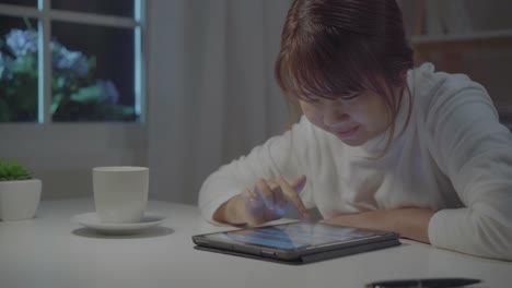 Junge-asiatische-Frau,-die-spät-mit-Tablet-Check-Social-Media,-während-entspannen-Sie-auf-dem-Schreibtisch-in-der-Nacht-im-Wohnzimmer-zu-Hause.-Genießen-Sie-die-Zeit-im-heimischen-Konzept.