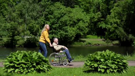 Lockdown-side-view-shot-of-senior-woman-pushing-her-disabled-husband-in-wheelchair-along-path-near-pond-in-green-park-on-warm-spring-day.-Mature-spouses-talking-cheerfully-and-enjoying-walk-in-nature