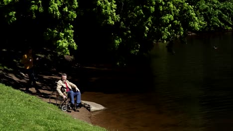 Frau-oder-Betreuer-bringen-Kaffee-für-behinderte-Männer-im-Rollstuhl-warten-am-See-im-Park.-Paar-beim-Kaffee-und-Gespräch-am-windigen-Sommertag