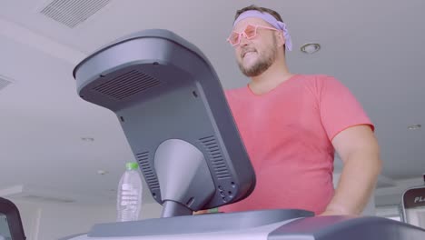 Funny-fat-male-in-pink-glasses-and-in-a-pink-t-shirt-is-engaged-on-a-treadmill-in-the-gym-depicting-a-girl.-4k