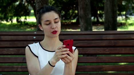 Close-up-portrait-A-beautiful-young-girl-in-a-white-T-shirt-is-chatting-in-social-networks-on-her-smartphone-while-sitting-on-a-bench-in-a-park.