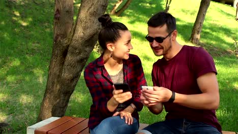 Couple-in-love-show-photos-on-their-smartphones-with-each-other,-sitting-on-a-park-bench.