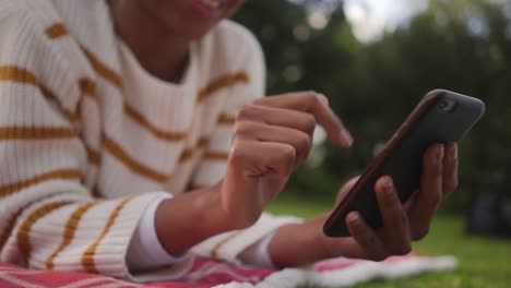 Primer-plano-de-una-joven-africana-que-yacía-en-el-parque-usando-la-aplicación-de-medios-sociales-en-el-teléfono-móvil