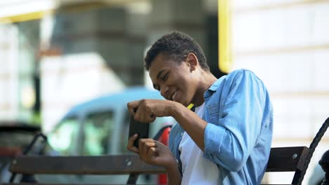 Excited-teenager-playing-action-game-on-smartphone-sitting-on-bench-,-addiction