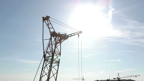 Two-construction-cranes-on-the-background-of-blue-sky-and-the-sun,-which-shines-in-the-camera-hiding-behind-the-construction-crane