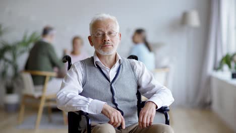 Tracking-medium-shot-of-disabled-elderly-man-in-eyeglasses-sitting-in-wheelchair-and-thinking,-then-turning-head-and-looking-at-camera-in-nursing-home,-other-aged-patients-in-background