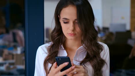 closeup-of-beautiful-young-business-woman-with-smartphone-texting-messenger-inside-office-in-the-background