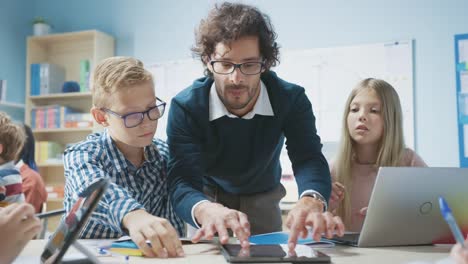 Grundschulklassenzimmer:-Enthusiastischer-Lehrer-mit-Tablet-Computer-erklärt-brillanten-kleinen-Kindern-den-Unterricht.-Kinder-lernen-Programmiersprachen,-Internetsicherheit-und-Softwaredesign