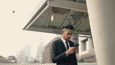 Schöne-junge-Geschäftsmann-mit-einem-Smartphone-SMS-Überprüfen-E-Mail-Nachrichten-online-auf-der-Straße-urban-mit-Himmel-Zug-auf-Hintergrund.