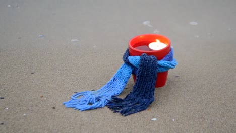 Red-mug-with-coffee-tied-with-blue-knitted-scarf-stands-on-sandy