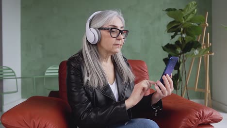 Front-view-of-beautiful-smiling-modern-60-aged-woman-with-long-gray-hair-which-wearing-glasses-and-enjoying-nice-music-on-headphones