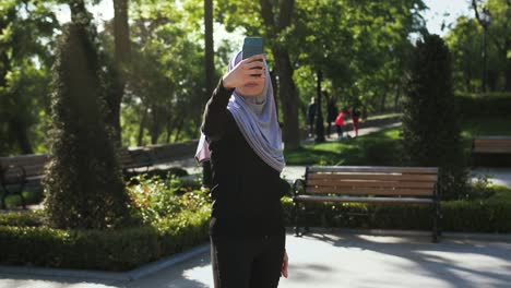 Attraktive-muslimische-Dame-in-lässigen-Kleidern-und-grauem-Hijab.-Sie-lächelt-und-macht-Selfie-auf-ihrem-Smartphone,-während-sie-in-einem-grünen-Landschaftspark-posiert