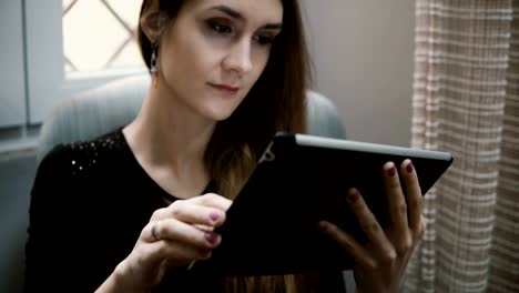 Young-attractive-woman-sitting-on-the-chair-and-chatting-on-social-networks,-using-tablet-computer-for-this