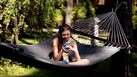 A-girl-in-a-blue-swimsuit-uses-her-phone-and-swings-in-a-hammock-in-nature.