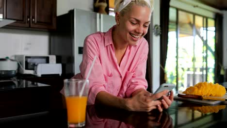 Mädchen-Einsatz-Zelle-Smart-Phone-innen-Küche-Studio-im-Chat-Online-junge-Frau-trinken-Saft-am-Morgen-modernes-Haus