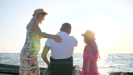 esposo-en-la-esposa-de-abrazo-de-silla-de-ruedas-con-gran-barriga,-familia-primer-plano-mirando-las-olas,-con-discapacidad-abraza-a-su-esposa-y-su-hija