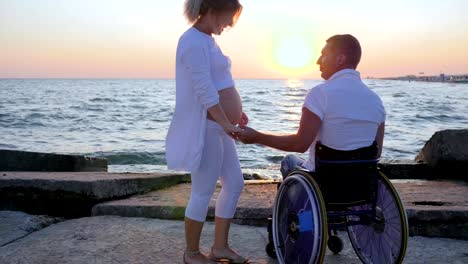 Handicapped-with-pregnant-girl-watching-sunset,-spouse-in-wheelchair-holds-arm-woman-with-big-belly,-happy-couple