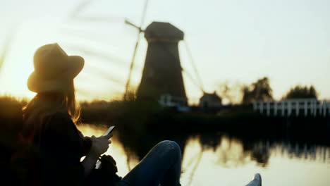 Chica-milenaria-se-encuentra-con-teléfono-en-el-muelle-del-lago.-Puesta-del-sol.-Mujer-de-uso-comercial-al-aire-libre-de-la-aplicación.-Molino-de-viento-holandés-antiguo.-4K