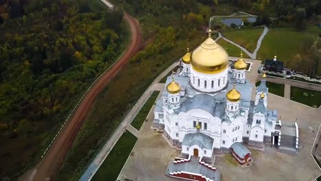 Aerial-view-on-great-white-christian-church-with-golden-domes-on-the-hill-and-field-with-forest-background.-Clip.-Top-view-on-Temple-in-the-middle-of-the-forest