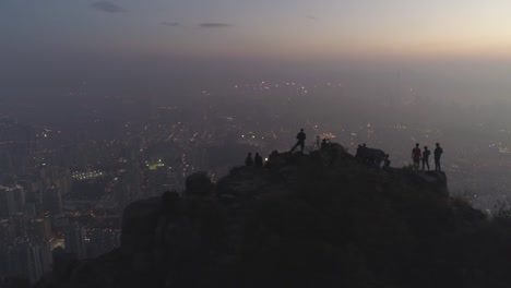 Silhouettes-of-people-on-the-top-of-rock-at-twilight.-Illuminated-City-Skyline-in-Smog.-Aerial-View.-Drone-is-Flying-Sideways.-Establishing-shot.