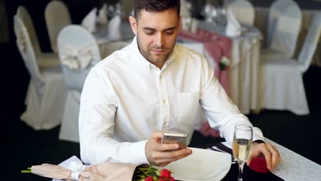 Novio-cariñoso-está-esperando-a-su-chica-en-el-restaurante,-con-smartphone,-apertura-de-caja-de-la-joyería-y-mirando-en-el-ring.-Ramo-de-rosas-y-Copa-de-champagne-son-accesibles.