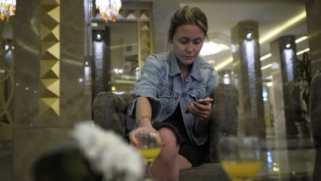 young-Caucasian-woman-in-a-jeans-jacket-drinks-juice-from-a-glass-and-uses-a-smartphone,-writes-messages-on-the-social-network.-Sits-at-the-table-in-the-hotel-restaurant.-Concept-of-healthy-fresh-food-in-business