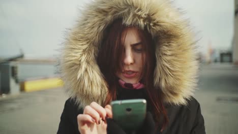 Close-up-shot-of-beautiful-woman-using-smart-phone-technology-app-walking-on-the-street-in-a-warm-jacket-with-a-fluffy-hood