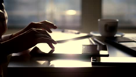 closeup,-dark-silhouette-of-female-hands.-she-is-typing-something-in-tablet,-next-is-a-cup,-mobile,-laptop-on-table.-Blurred-window-background-,-a-ray-of-light-is-reflected-in-the-tablet-screen