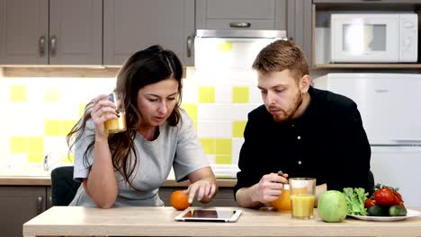 Young-couple-man-and-woman-with-tablet-eating-breakfast-sitting-by-table-in-kitchen-at-home.